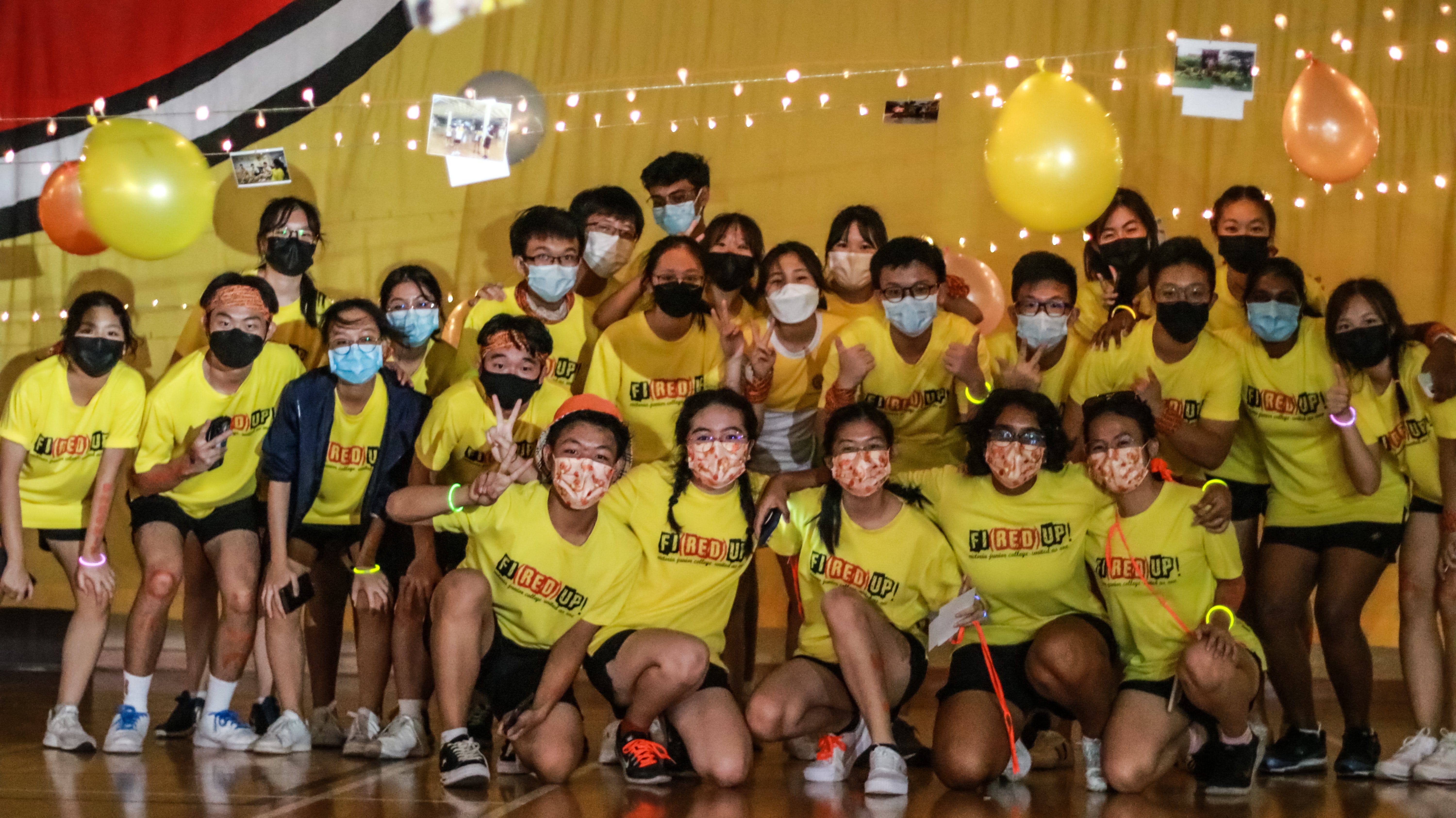 Large group of masked students in yellow "Fired Up" shirts pose together in a decorated gym with balloons and string lights.